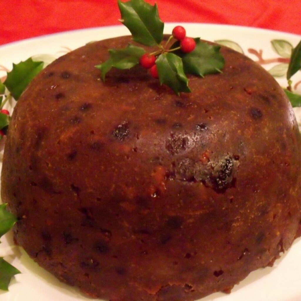 A brown Christmas pudding on a platter with holly on top as a decoration