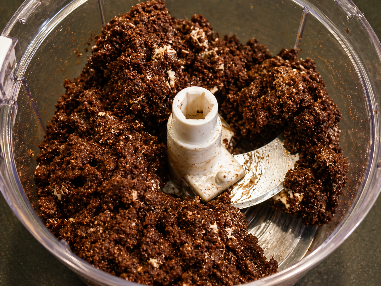 Chocolate cookie dough crumbs in a white plastic food processor with a central blade guard.