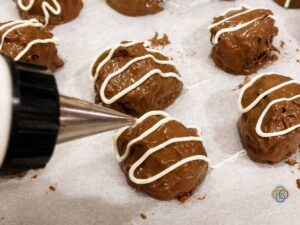 Chocolate-coated truffles on parchment being drizzled with white chocolate from a piping bag.