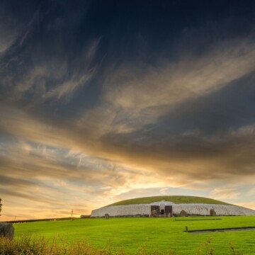 The sun illuminates a large stone burial mound in a green field