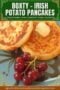 Two golden Irish potato pancakes (boxty) served on a plate with a pat of butter melting on top and a small bunch of red grapes on the side with green and white toned text banner.