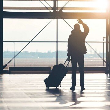 A silhouette of a man waving at the airport while holding a bag on wheels