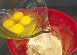 Four eggs in a pitcher being poured into flour in a bowl
