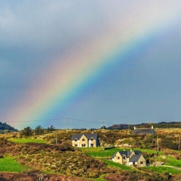 A rainbow arc over green and rocky fields and two homes