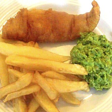 A white plate with a piece of beer battered fish served with chips or french fries and green mushy peas