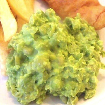 Green mushy peas on a plate with a few chips and the end of a piece of battered fish in the background