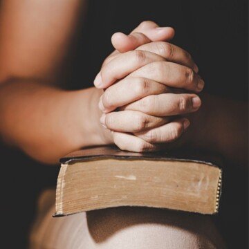 Hands held in prayer over a Bible balanced on a person's knee