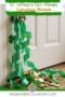 St. Patrick’s Day leprechaun blockade prank with green ribbon hanging from a door handle, shamrocks and gold coins scattered on the floor, featuring Irish American Mom branding.