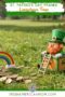 Toy leprechaun sitting in a wooden trap box with ladder beside gold coins and a rainbow on grass, labeled “St. Patrick’s Day Pranks Leprechaun Traps” with Irish American Mom branding.