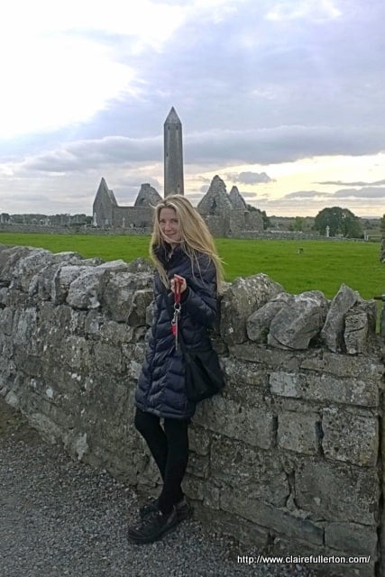 Claire Fullerton | Irish American Mom A person standing in front of a stone wall with a round tower and ruined church in the background