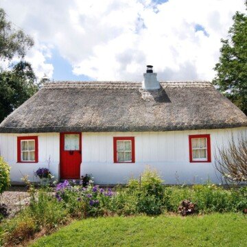 A white cottage with red door and windows with red frames and a thatched roof