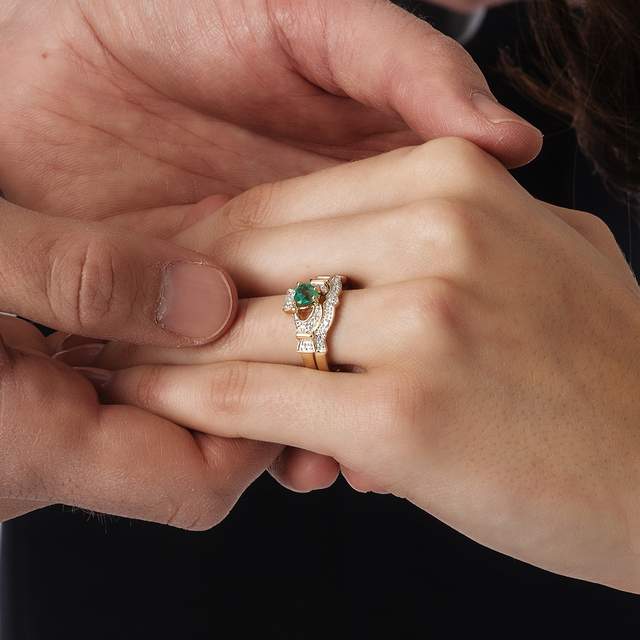 A man holding a woman${2}s left hand with a Claddagh emerald and diamond engagement ring