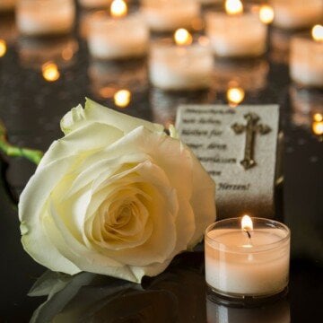 A white rose, a candle and a religious plaque on a black marble surface in front of rows of lighting candles