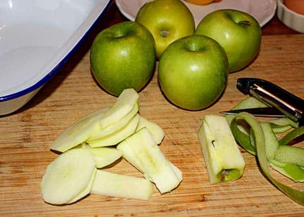 Peeled, sliced and cored apples for Eve's Pudding | Irish American Mom Green apples beside a sliced and cored apple with peels and a peeler on a chopping board.