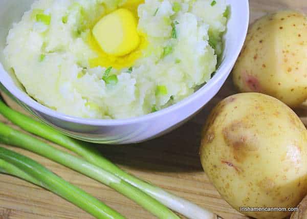 A bowl of Irish champ - mashed potatoes mixed with green onions and milk | Irish American Mom A bowl of mashed potato champ with melting butter beside potatoes and green onions