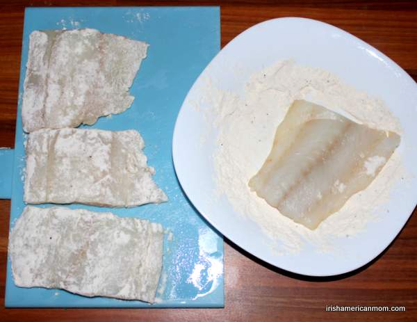 Dredging cod in seasoned flour for cod and parsley sauce | Irish American Mom Cod fillets on a blue chopping board with one piece being dredged in flour in a white bowl.