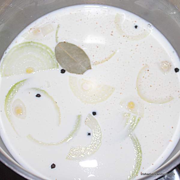 Simmering milk with onion, peppercorns, bay leaf and nutmeg for parsley sauce | Irish American Mom Milk in a saucepan with onion slices, peppercorns and bay leaf