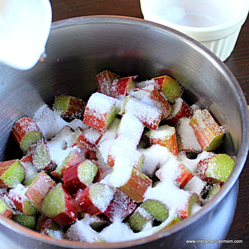 Adding sugar and water to rhubarb pieces in a saucepan to parboil it for rhubarb crumble | Irish American Mom Sugar being sprinkled over chopped rhubarb in a silver pot