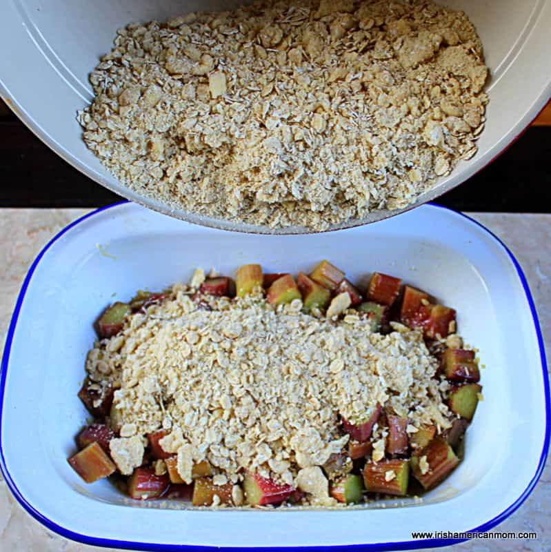 Covering parboiled rhubarb with crumble topping to make Irish rhubarb crumble | Irish American Mom Pouring an oaty crumble mixture over rhubarb in a casserole