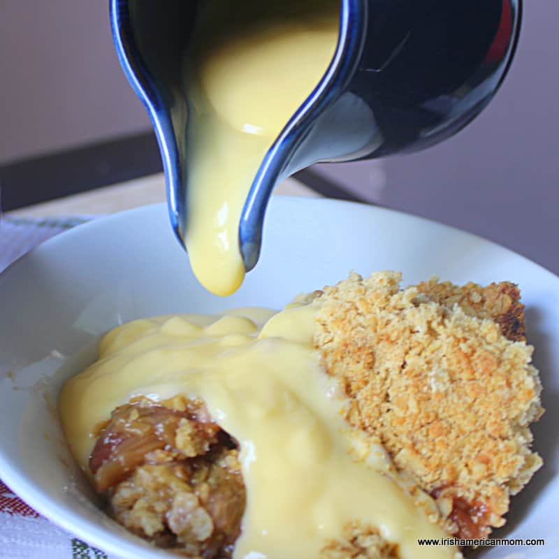 Pouring hot custard over a serving of rhubarb crumble in a bowl | Irish American Mom Custard dripping from a pitcher over a serving of rhubarb crumble in a bowl.