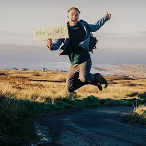 Hitching for Hope Irish author Ruairi Mc Kiernan with a hiking sign | Irish American Mom A man jumps in the air holding a sign saying hitching for hope.