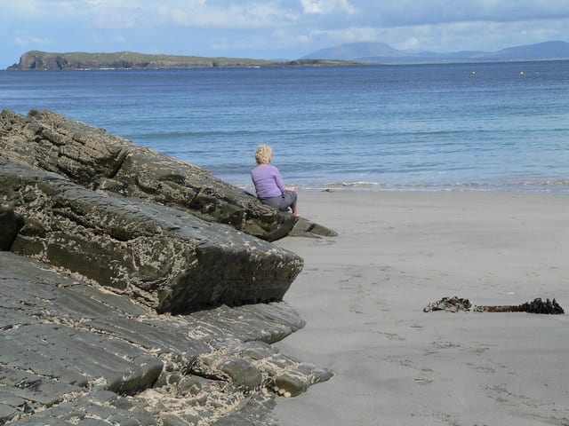 A blonde woman in a purple sweater sits on a rock by a sandy beach looking out to sea at a distant island