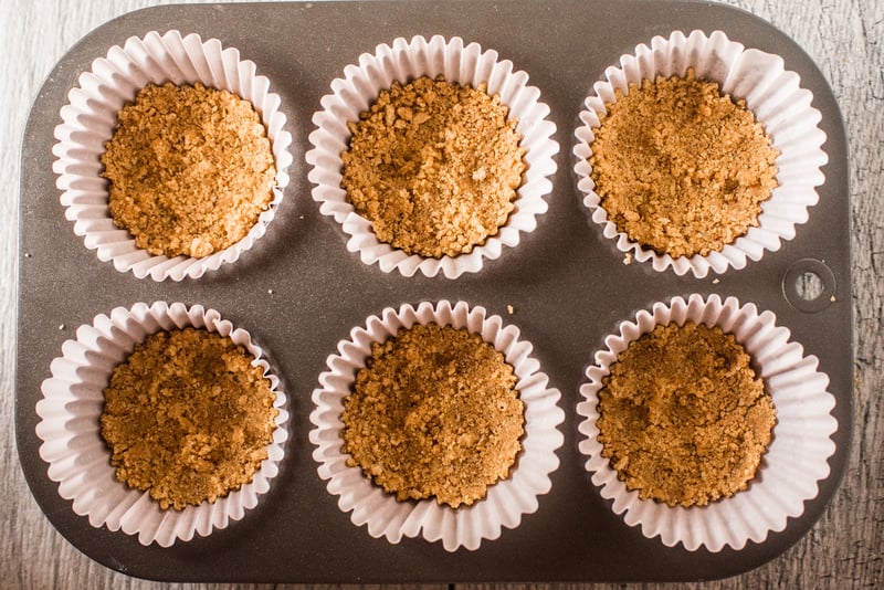 A six place muffin pan with paper lines with a layer of graham cracker crumbs at the base