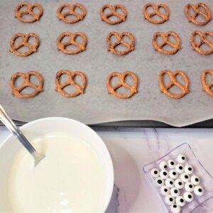 A bowl of melted white chocolate beside pretzels on a baking tray and a container of candy eyes