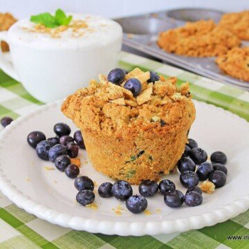 A blueberry muffin with a crunchy topping on a white plate beside a creamy coffee in a white cup