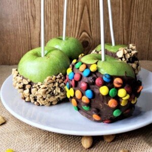 Four green apples half coated in chocolate and two dipped in candies and two dipped in crushed nuts