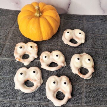 A small orange pumpkin beside a selection of ghost chocolate pretzels with candy eyes