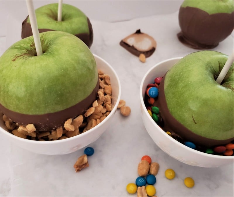 Chocolate coated apples being dipped in crushed peanuts and chocolate button candies | Irish American Mom Green apples dipped in melted chocolate being coated in nuts and in candies in two white bowls