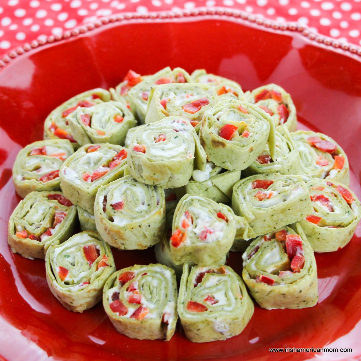 A red plate with a stack of festive pinwheel sandwiches with red peppers speckled through the filling
