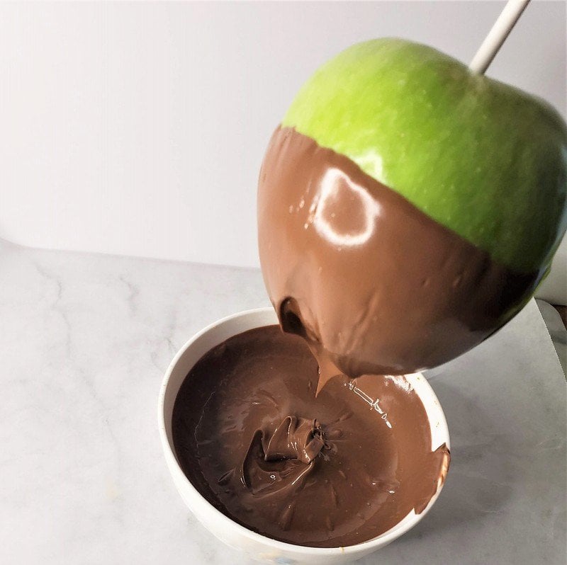 Coating the lower half of an apple in melted chocolate | Irish American Mom Lifting a green apple out of a bowl of melted chocolate after coating half the apple in the chocolate