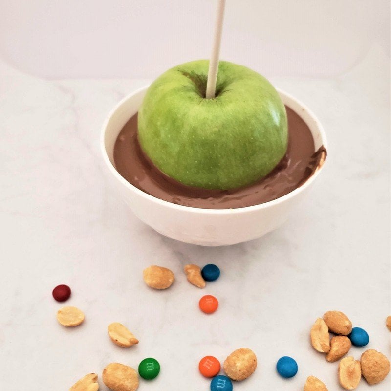 Dipping a green apple in melted chocolate | Irish American Mom A green apple on a lollipop stick being dipped into a white bowl of melted chocolate