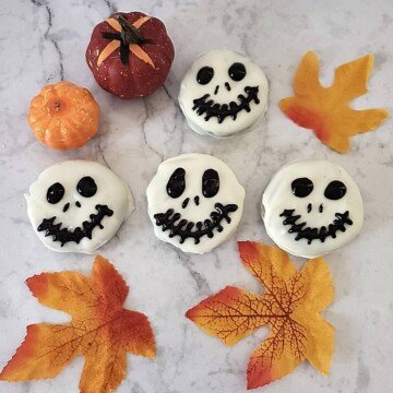 Black skeleton faces on white chocolate dipped cookies displayed beside fall leaves and miniature decorative pumpkins