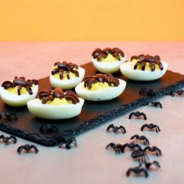 Deviled eggs with spider decorations made out of olives served on a black slate with an orange Halloween backdrop
