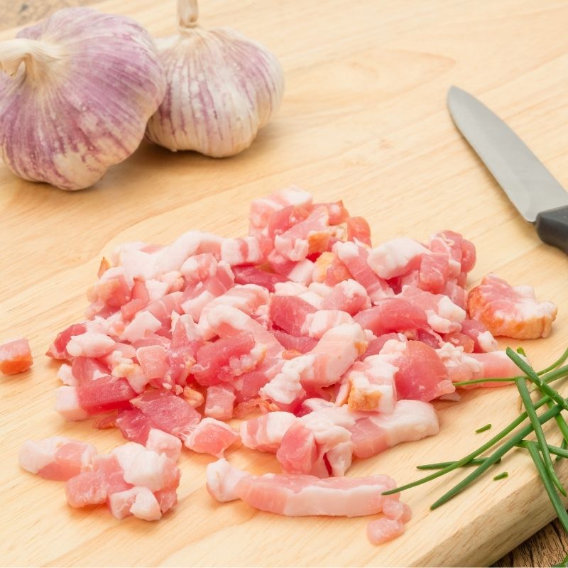 Bacon lardons on a chopping board | Irish American Mom A wooden chopping board with chopped bacon pieces beside a knife and two garlic bulbs
