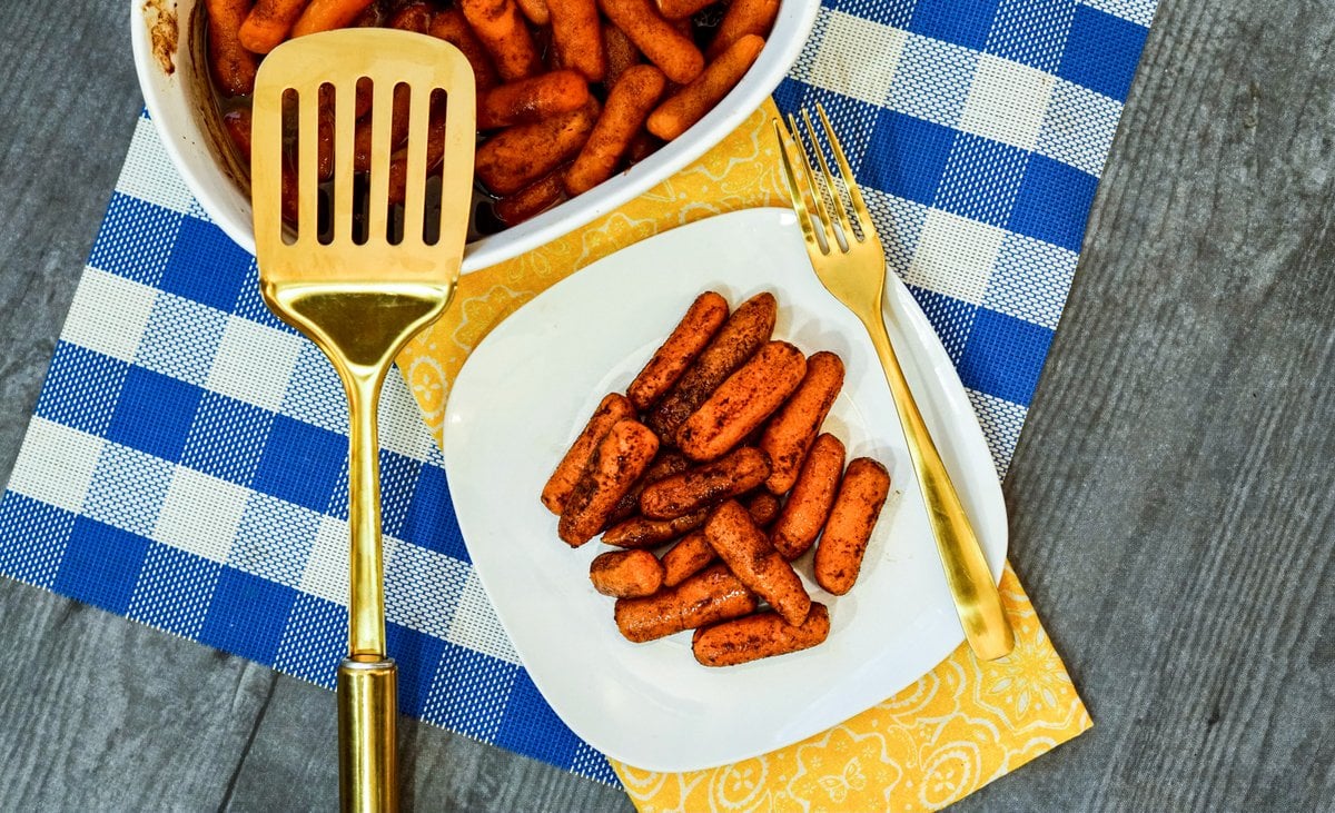 Baked and glazed carrots on a plate beside a casserole dish of carrots | Irish American Mom Carrots on a plate with golden silverware and spatula on a blue check place mat