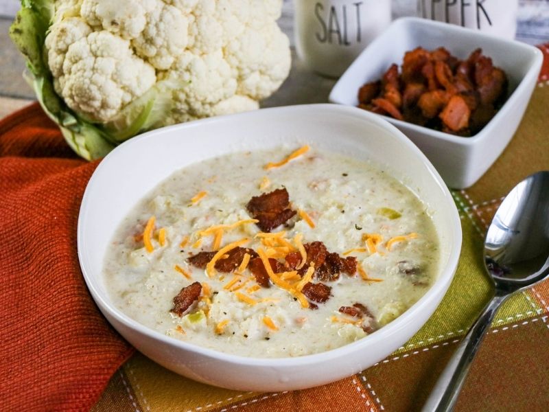 A bowl of cauliflower soup with bacon bits displayed beside a head of cauliflower