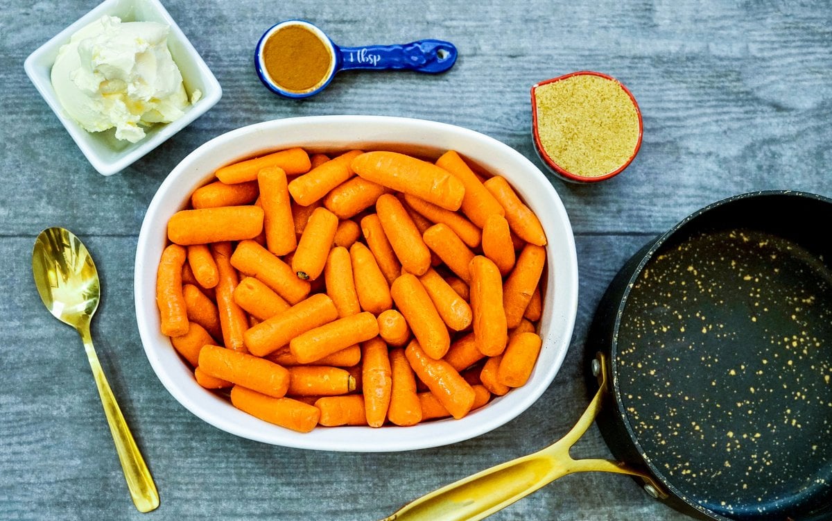 Ingredients for sugar glazed carrots | Irish American Mom Bowls of butter, carrots, brown sugar and a spoon of cinnamon beside a black skillet