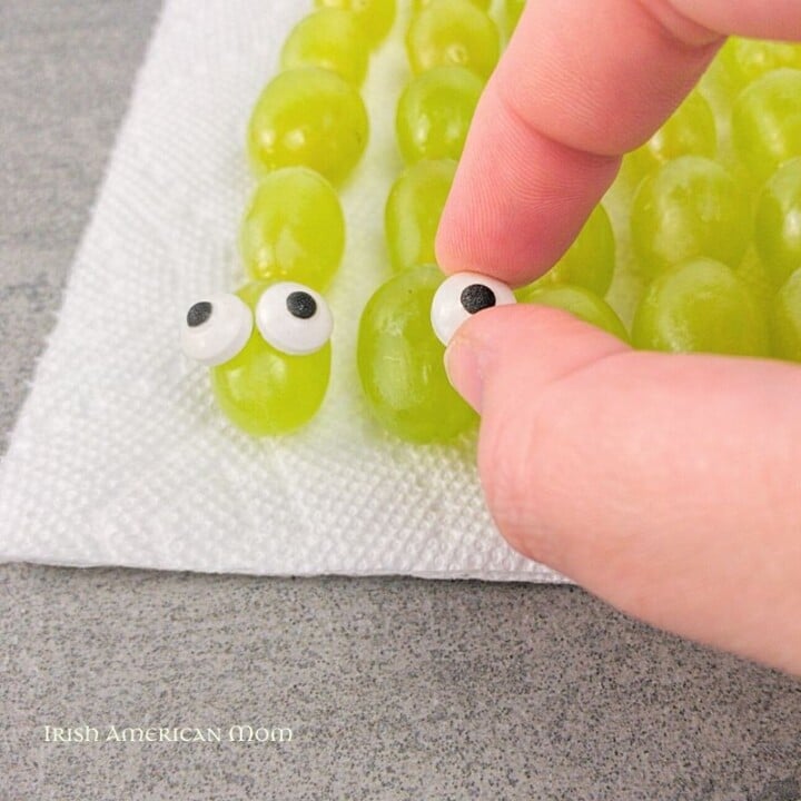 Adding candy eyes to grape snake snacks for Saint Patrick's Day | Irish American Mom Fingers placing candy eyes on green grape snakes