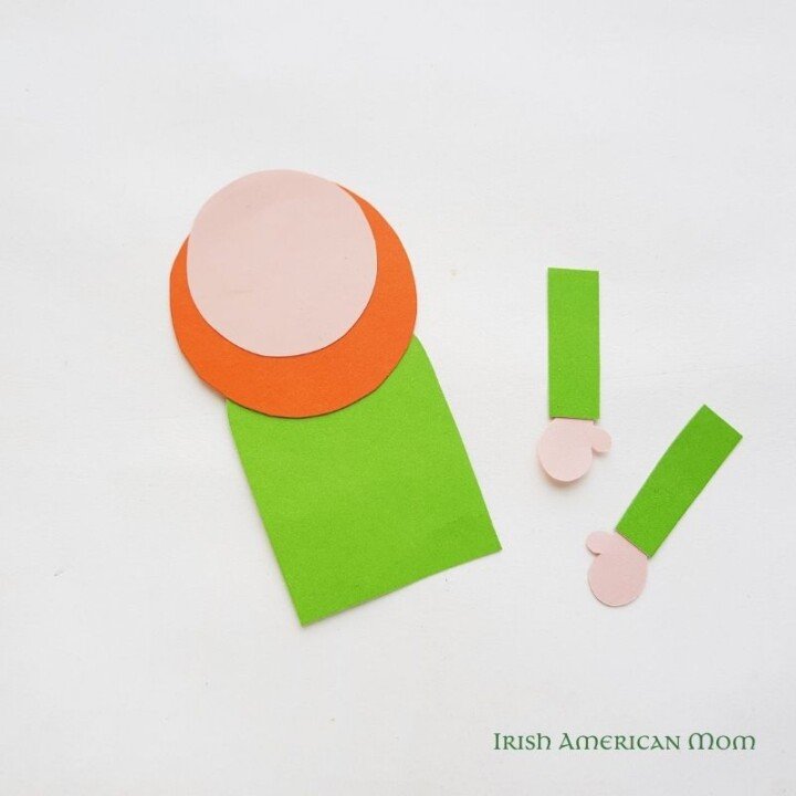 Gluing the leprechaun head to the body for a bookmark | Irish American Mom Pink and orange paper circles on to pf a green paper rectangle beside paper arms with pink hands