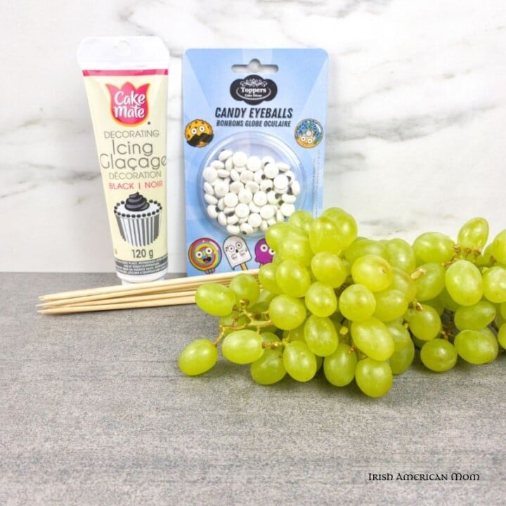 Ingredients for green grape snake snacks for Paddy's Day | Irish American Mom Icing tube, packet of candy eyes, green grape bunch and wooden skewers on a counter top