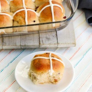 Yeast bun with an iced cross on a plate beside a dish of hot cross buns