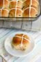 Single homemade hot cross bun with icing cross on a white plate, with a tray of baked buns in the background on a cloth surface.