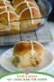 Homemade Irish hot cross bun with icing cross on a white plate, with a baking dish of buns in the background on a rustic surface.