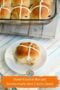 Homemade hot cross bun with icing cross on a white plate, with a glass baking dish of buns in the background on a patterned cloth.