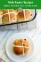 Homemade hot cross bun with icing cross on a white plate, with a glass dish of buns in the background on a patterned cloth surface.