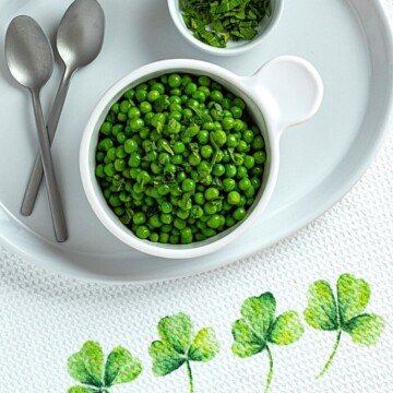 Peas in a bowl beside mint leaves, spoons and a shamrock dish cloth